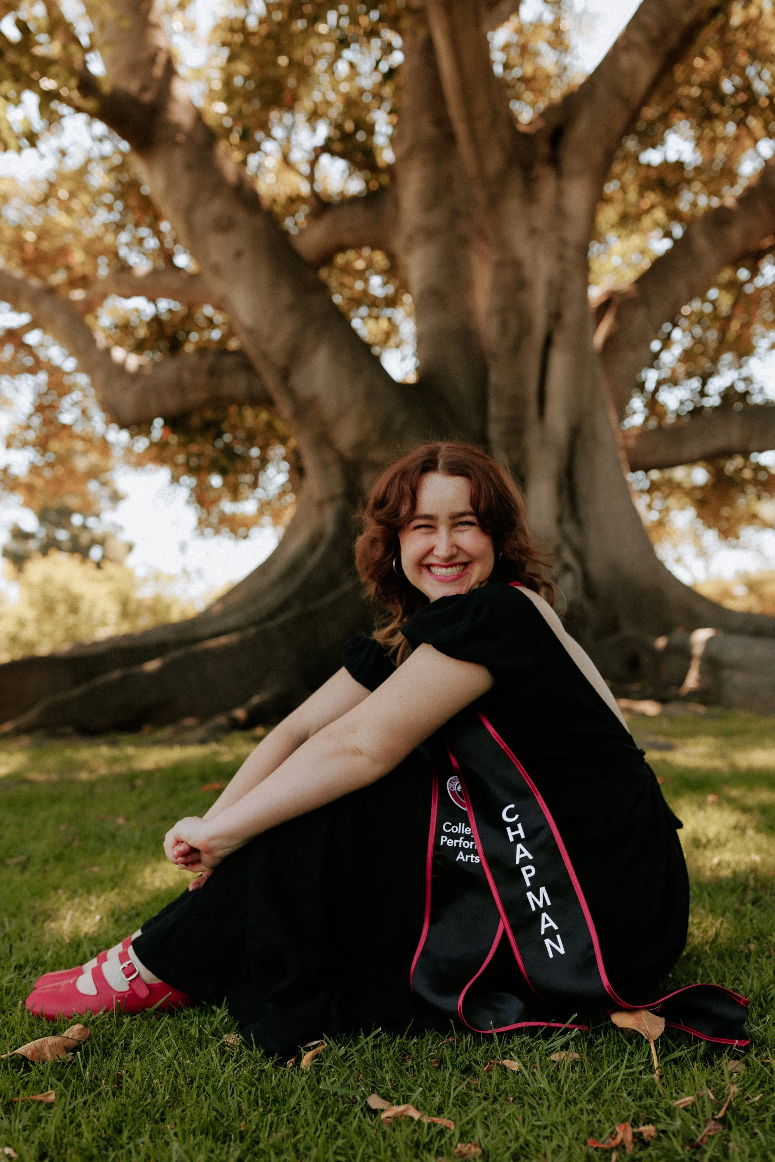 A young woman with curly red hair smiling and sitting on the grass in front of a large tree with autumn-colored leaves. She is wearing a black graduation gown with a sash that has the words 'Chapelman' and 'College Performance Arts' on it.
