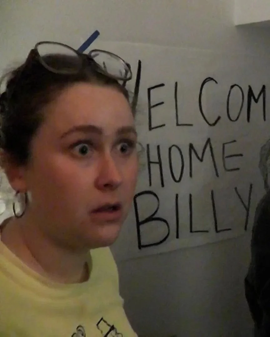 A woman with glasses on her head looking surprised in front of a sign that says 'Welcome Home Billy'.