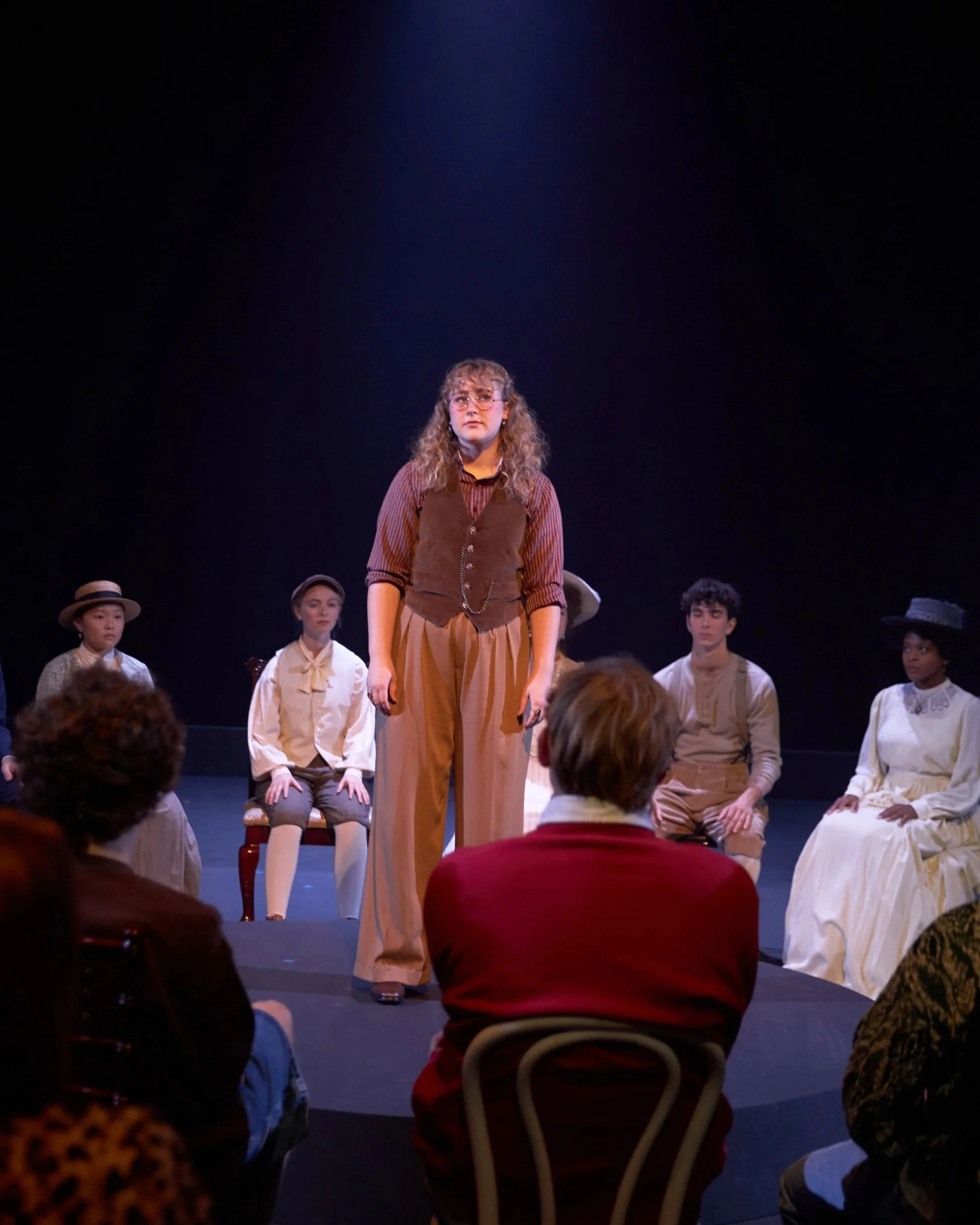 Stage play scene with a woman in the center dressed in vintage style clothing, with a dark background, and seated actors around her in period costumes.