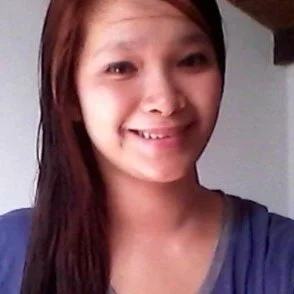 A woman smiling with long dark hair, wearing a blue top, indoors.
