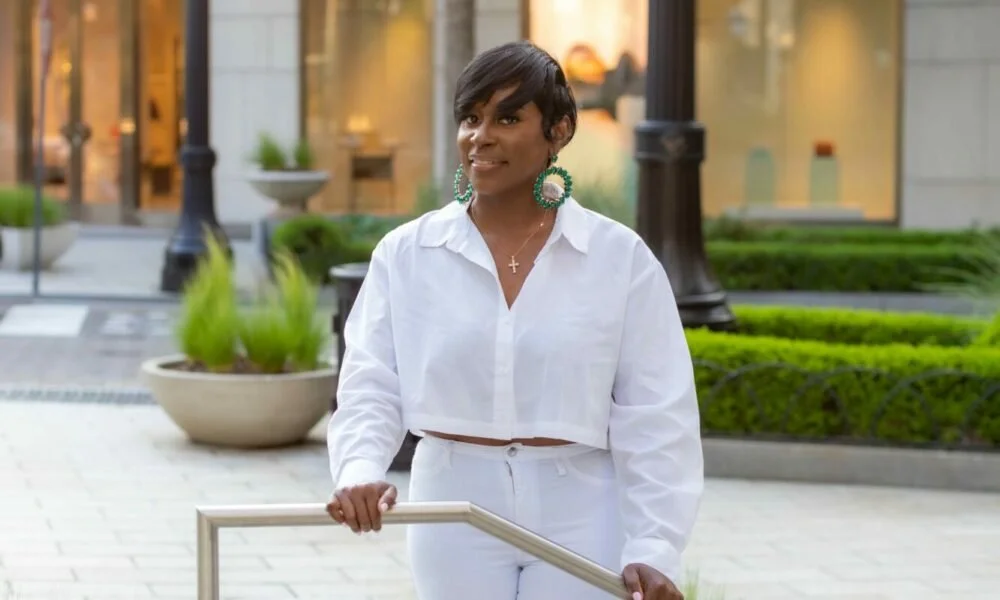 Woman with short dark hair, large hoop earrings, and a cross necklace, dressed in a white shirt and white pants, standing outdoors in an urban setting with buildings and greenery in the background.