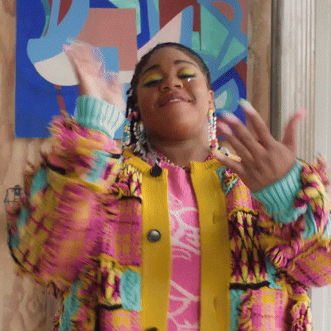 A girl with colorful makeup and large earrings, wearing a vibrant multicolor jacket, smiling and gesturing with her hands, standing in front of an abstract, colorful background.