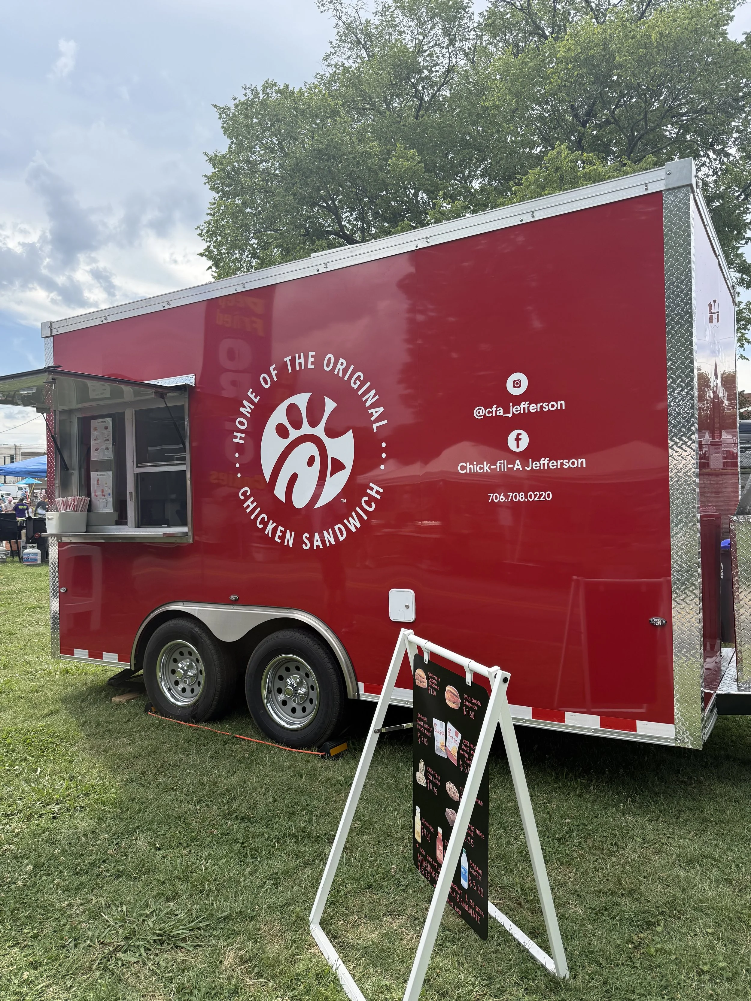 Red chicken sandwich food truck with a white logo and social media handles, standing on grassy area with trees and cloudy sky and people in the background.