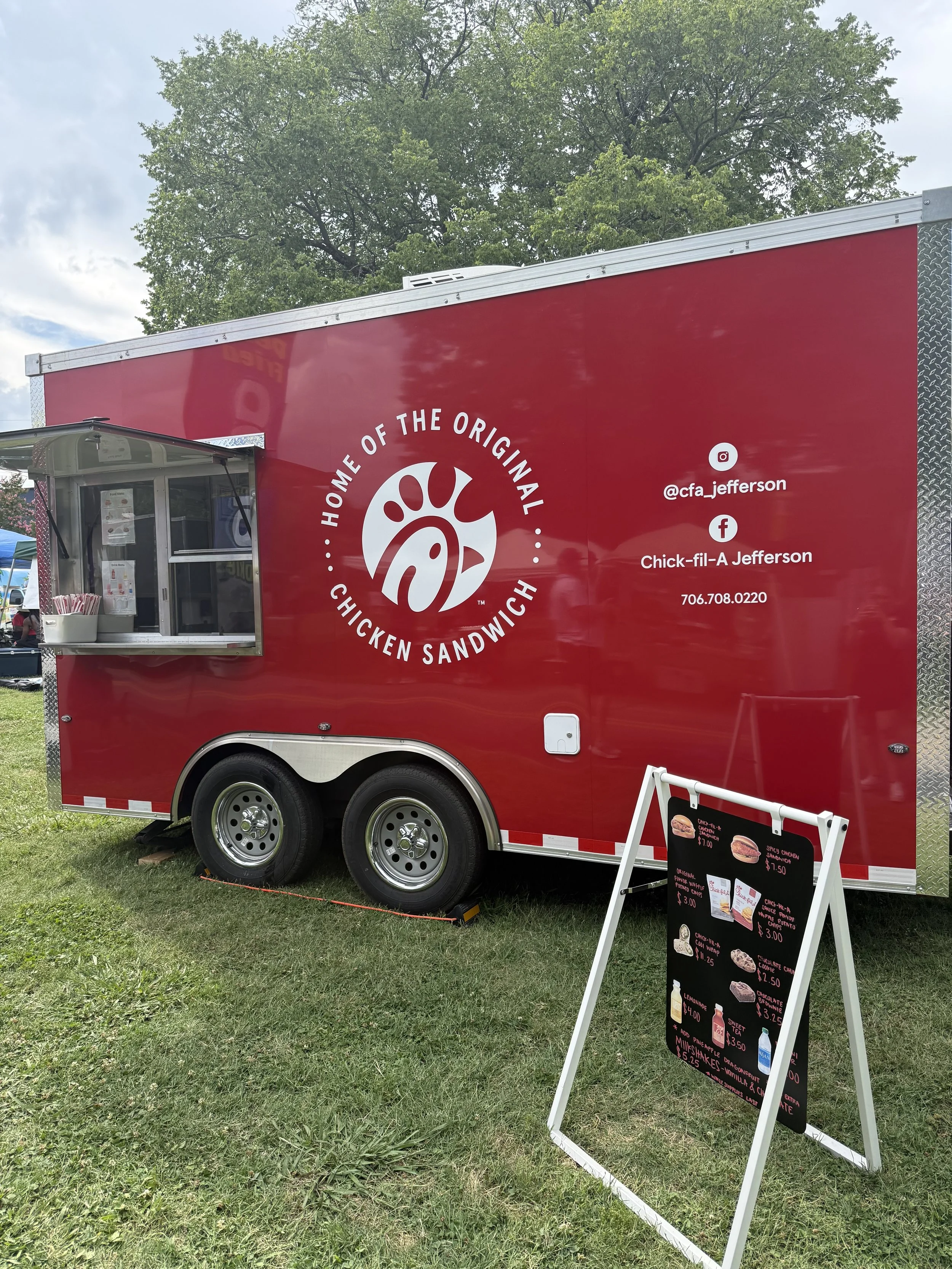 A red Chick-fil-A food truck with a menu board on a grassy area, featuring various chicken sandwiches, combos, and drinks, with trees and a partly cloudy sky in the background.