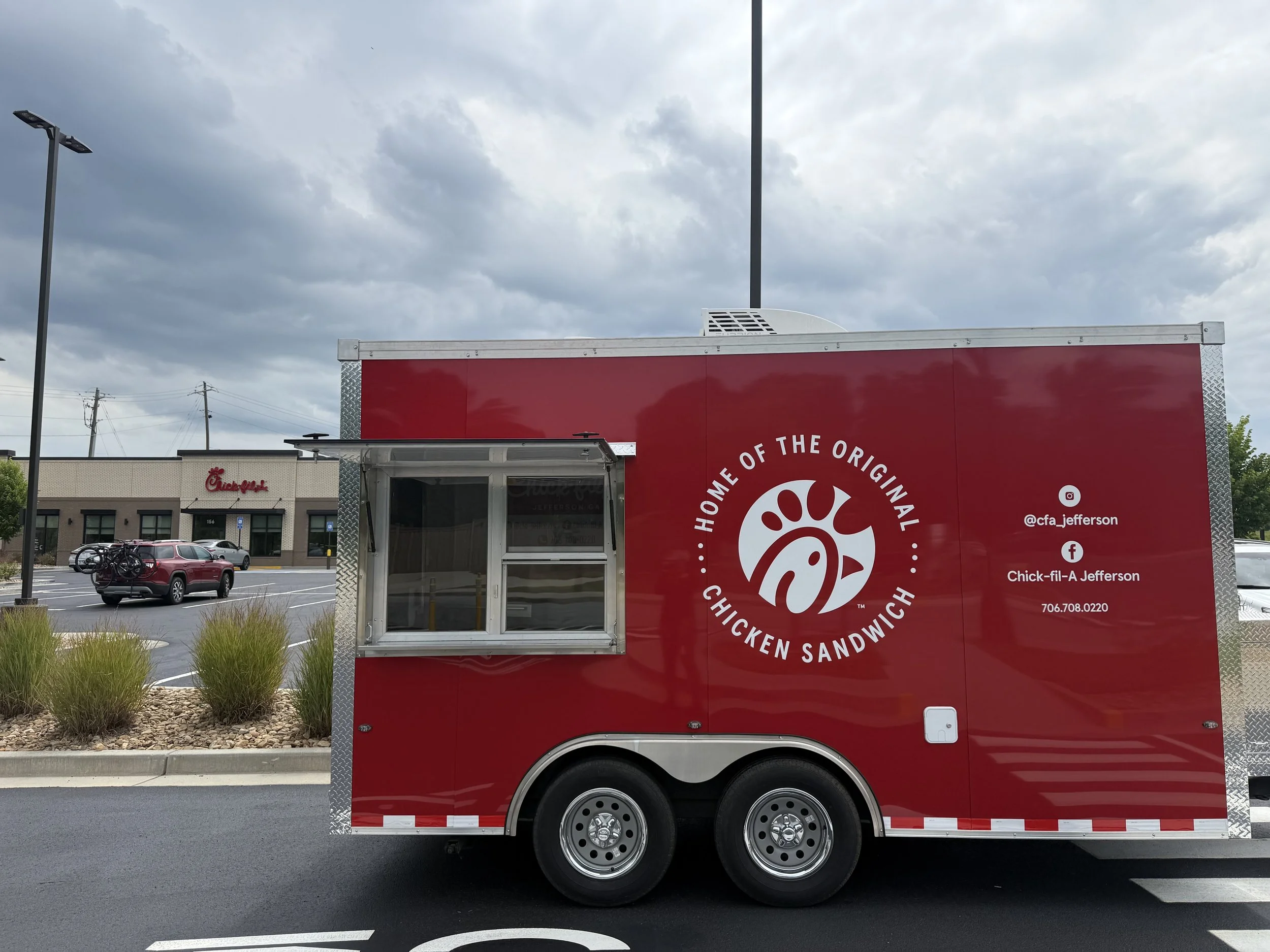 Red Chick-fil-A food truck parked in a lot with a Chick-fil-A restaurant and a parking lot with cars in the background. The truck has the Chick-fil-A logo and the words "Home of the Original Chick-fil-A Chicken Sandwich." The sky is cloudy.