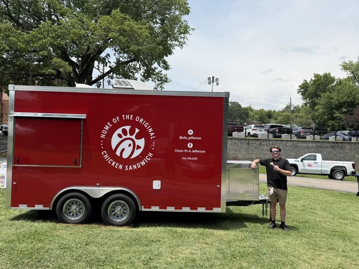 A man standing next to a red Chick-fil-A food truck on a grassy area, holding a smartphone and wearing sunglasses.