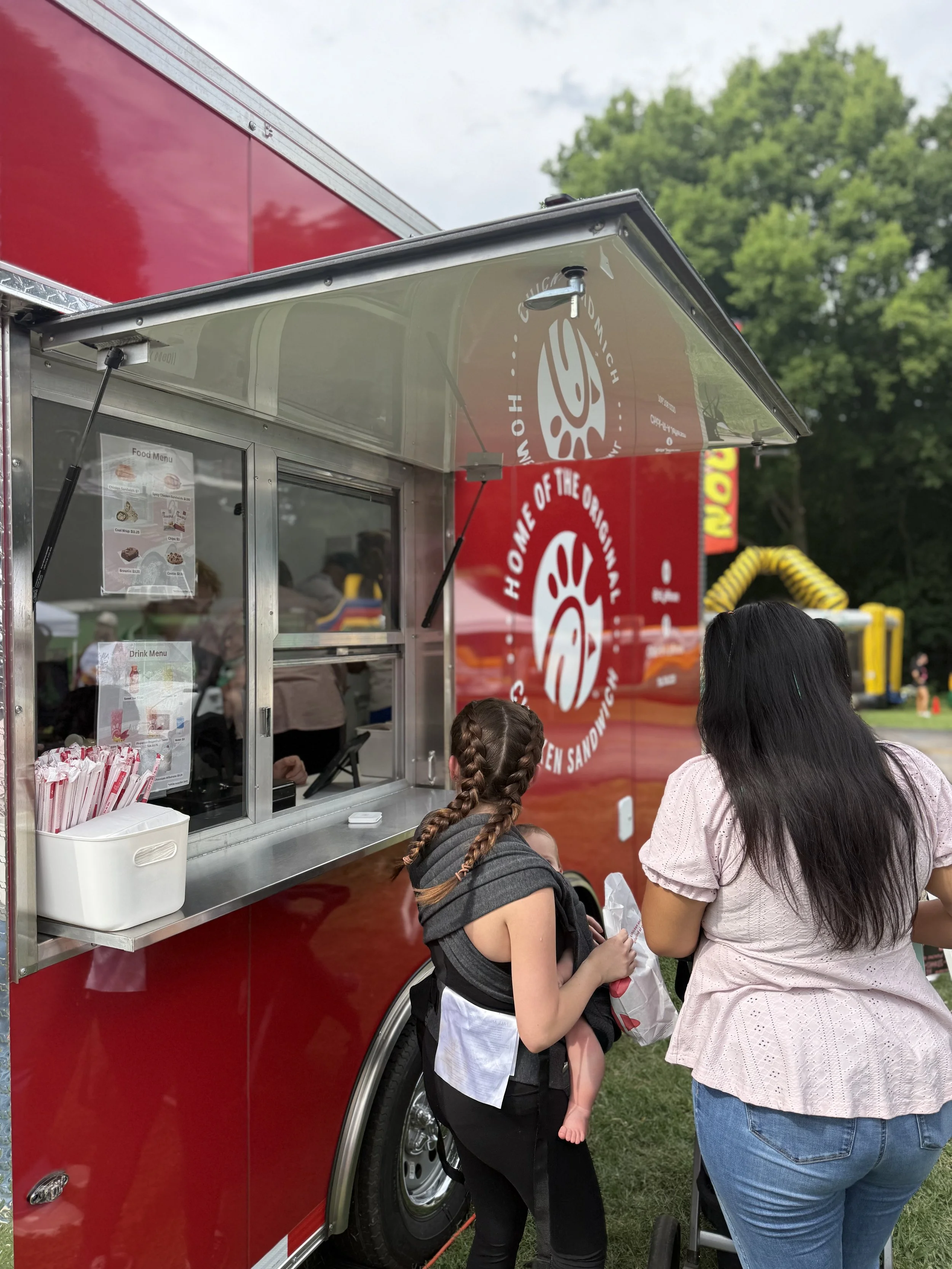 A red food trailer with a window serving area, where a woman and a girl are ordering food. The trailer has a logo that reads 'Home of the Original' with a smiling face image. The woman has long dark hair and is wearing a pink shirt. The girl with braided hair is wearing a black and white outfit. They are outdoors on green grass with trees and a yellow playground slide in the background.