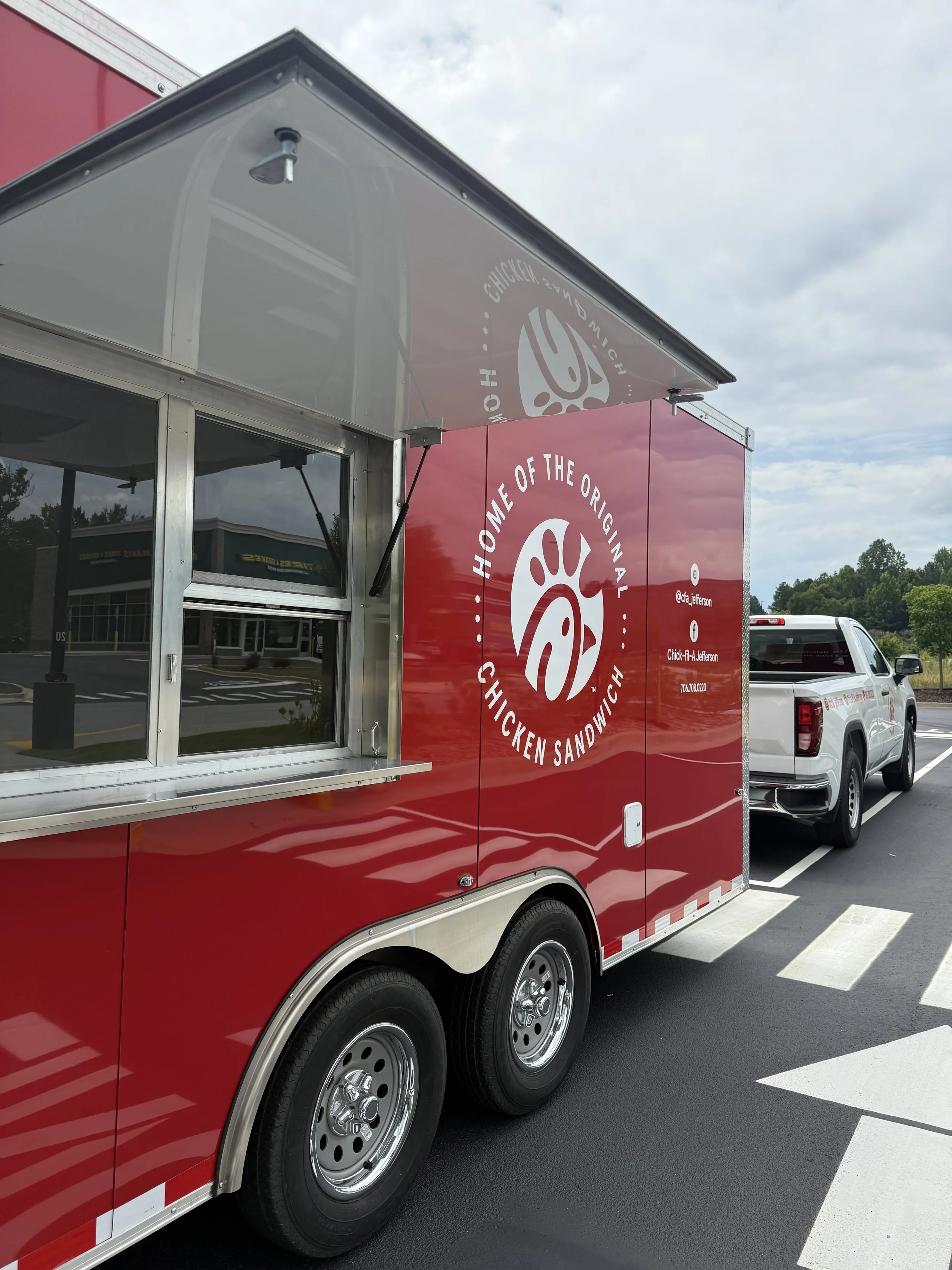 Red Chick-fil-A food truck with a serving window and logo, parked in parking lot with a white truck nearby, under a partly cloudy sky.