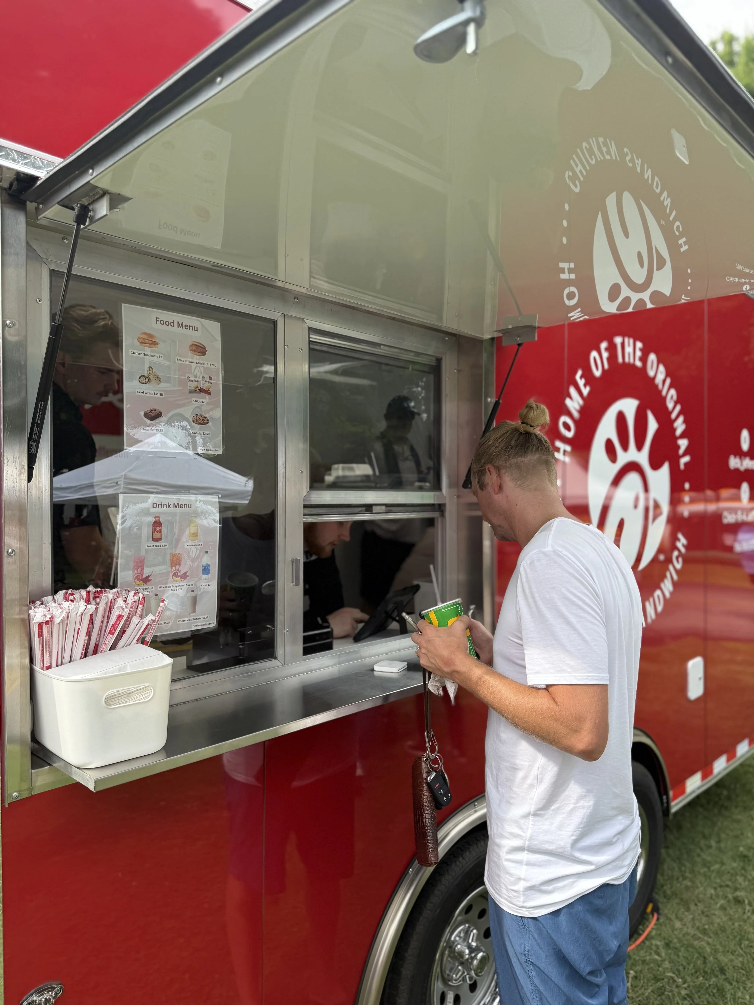 A man in a white t-shirt and blue jeans standing at a red food truck ordering food. The truck has a food menu posted on it and a container of straws. The man is holding a shop or snack package and has keys hanging from his pocket. The food truck has a logo and text that reads 'Home of the Original Chicken.'