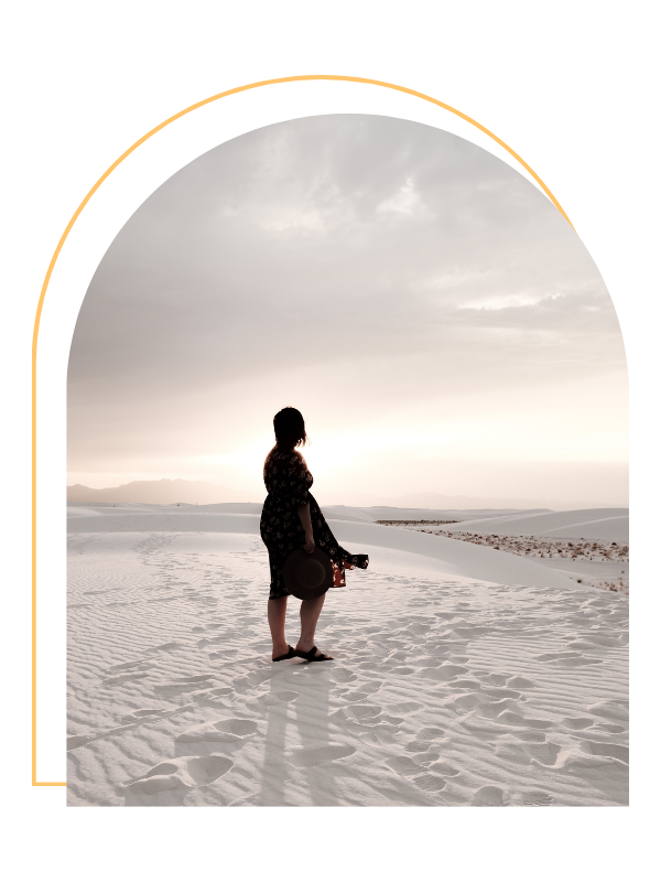 A woman standing alone in a vast desert with white sand dunes, holding a hat and looking towards the horizon at sunset or sunrise.