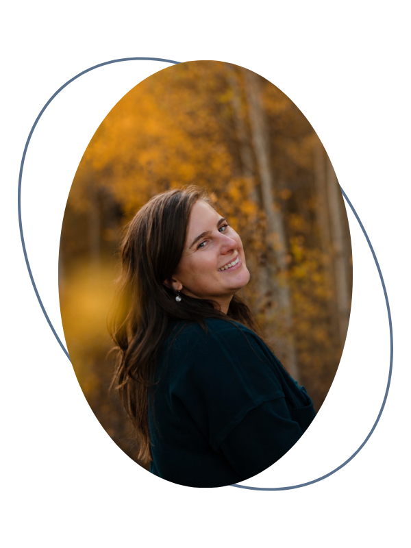 A woman with long brown hair smiling outdoors during fall, with yellow and orange foliage in the background.