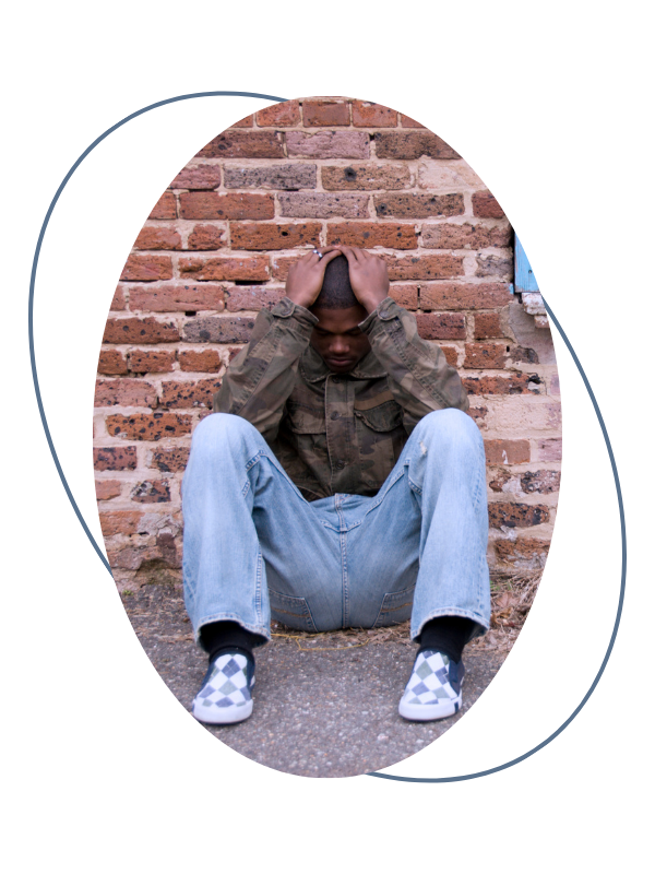 Young man sitting on the sidewalk against a brick wall, holding his head with both hands, appearing distressed.
