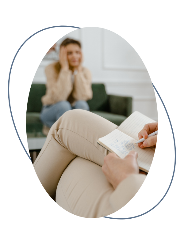 A woman sitting on a green couch with a distressed look, hands on her face, during a therapy session with a counselor. The counselor is taking notes in a notepad.