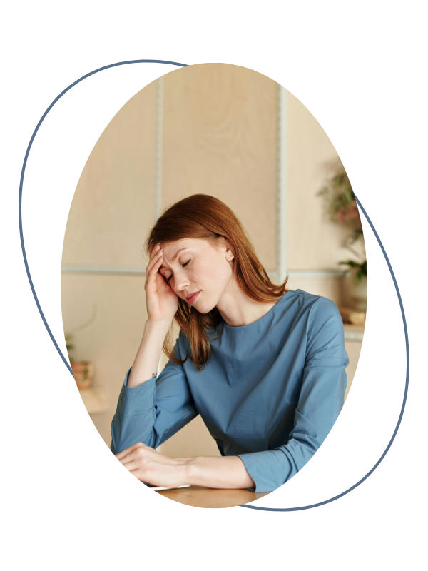 A woman with red hair holding her head in a thoughtful pose, sitting at a desk in an office or home setting.