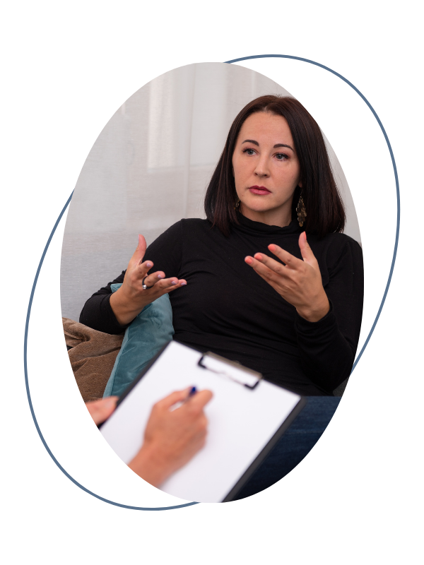 A woman with dark brown hair and a black turtleneck is sitting and gesturing with her hands during a conversation, while a person in the foreground is taking notes on a clipboard.