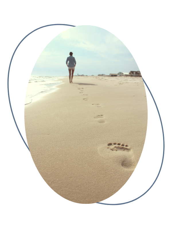 A person walking along a sandy beach with footprints in the sand leading towards the person, under a partly cloudy sky.
