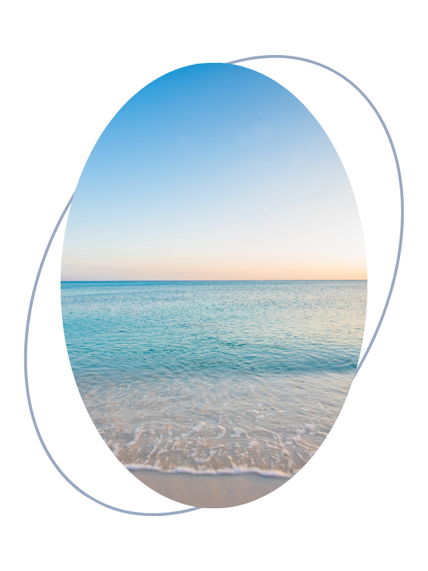 View of calm ocean water meeting sandy beach at sunset or sunrise, with clear sky and horizon.