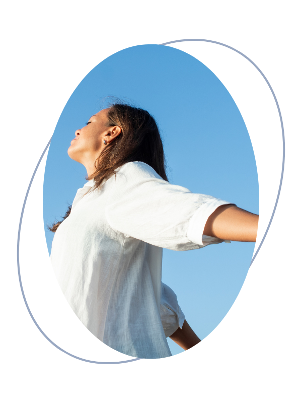 A woman with brown hair and earrings is stretching outdoors with her arms outstretched, facing the blue sky with her eyes closed in a relaxed pose.