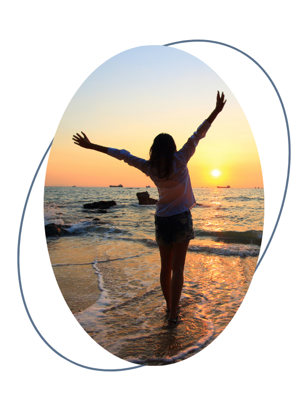 A woman standing in the water at the beach with her arms raised during a sunset.