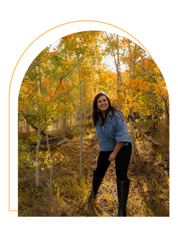 A woman with long dark hair smiling and posing in a forest during autumn, surrounded by colorful fall foliage, wearing a denim jacket, black pants, and tall black boots.