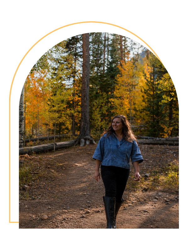 A woman walking on a dirt trail in a forest with trees showing fall foliage, wearing a denim jacket and black pants.