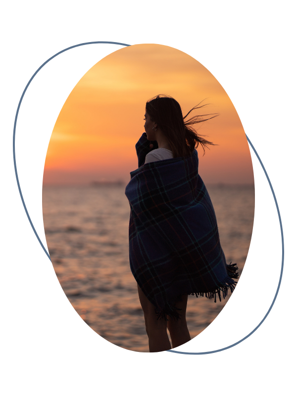 A woman wrapped in a plaid blanket standing by the water during sunset, looking out at the horizon.