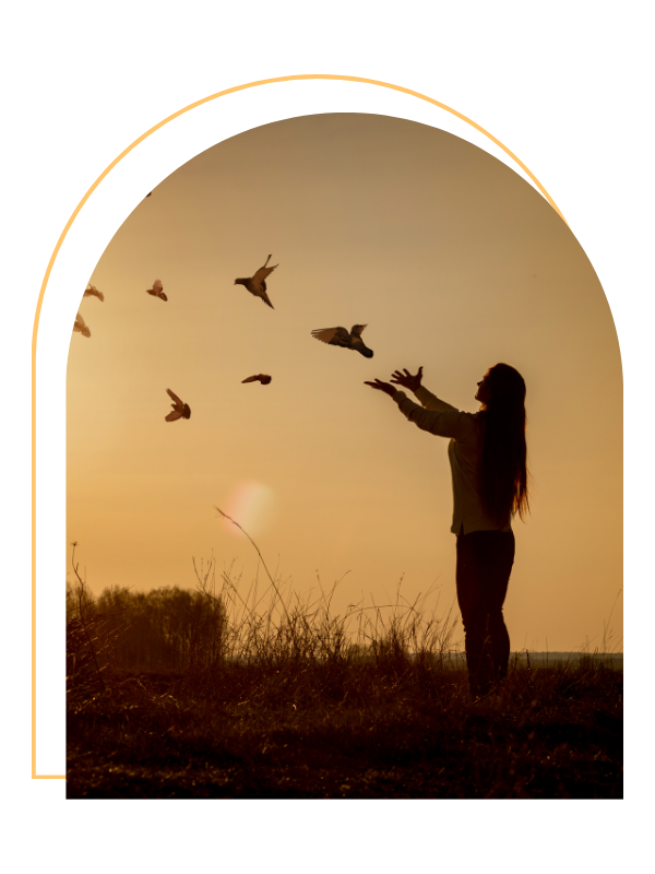 A woman stands outdoors during sunset, releasing birds into the sky.