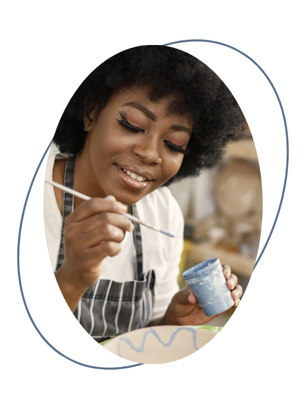 A woman with curly hair painting a ceramic object with blue glaze in a workshop setting.