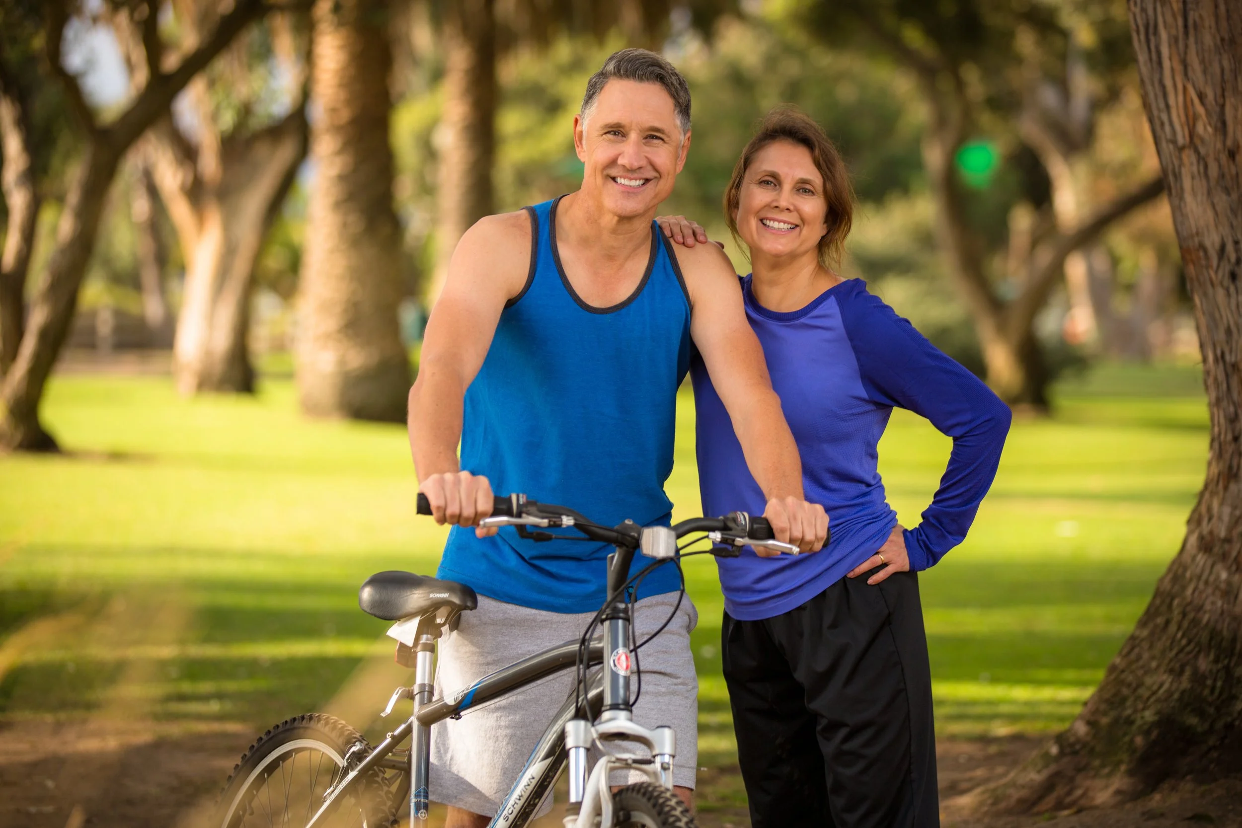 A middle-aged man with gray hair and a woman with brown hair standing close together in a park, smiling. The man is beside a bicycle with his hand on the handlebar, and the woman has her arm around the man's shoulder. They are wearing athletic clothes, and there are trees and greenery in the background.
