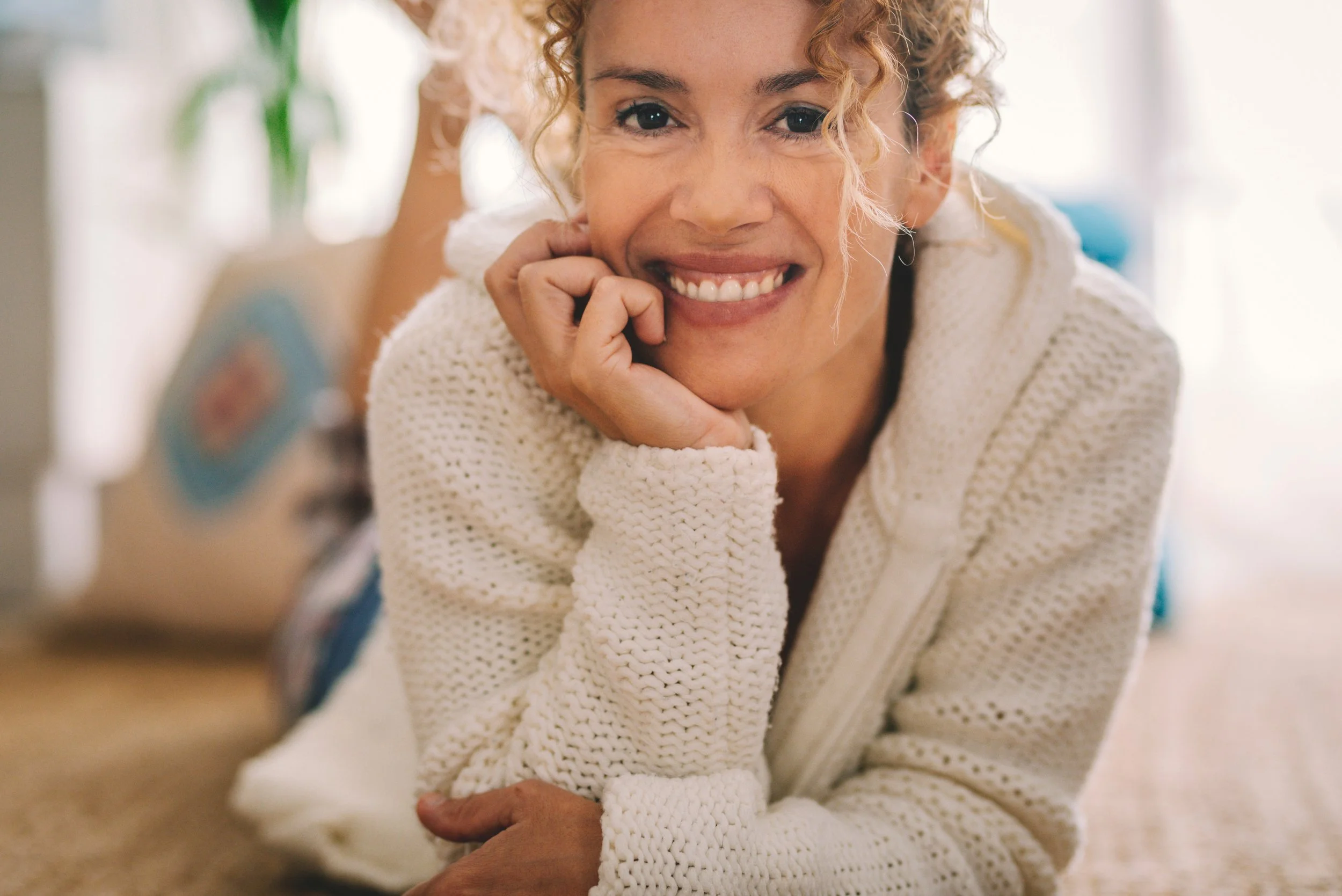 Close-up of a woman with curly hair smiling and resting her chin on her hand, wearing a white knitted sweater.