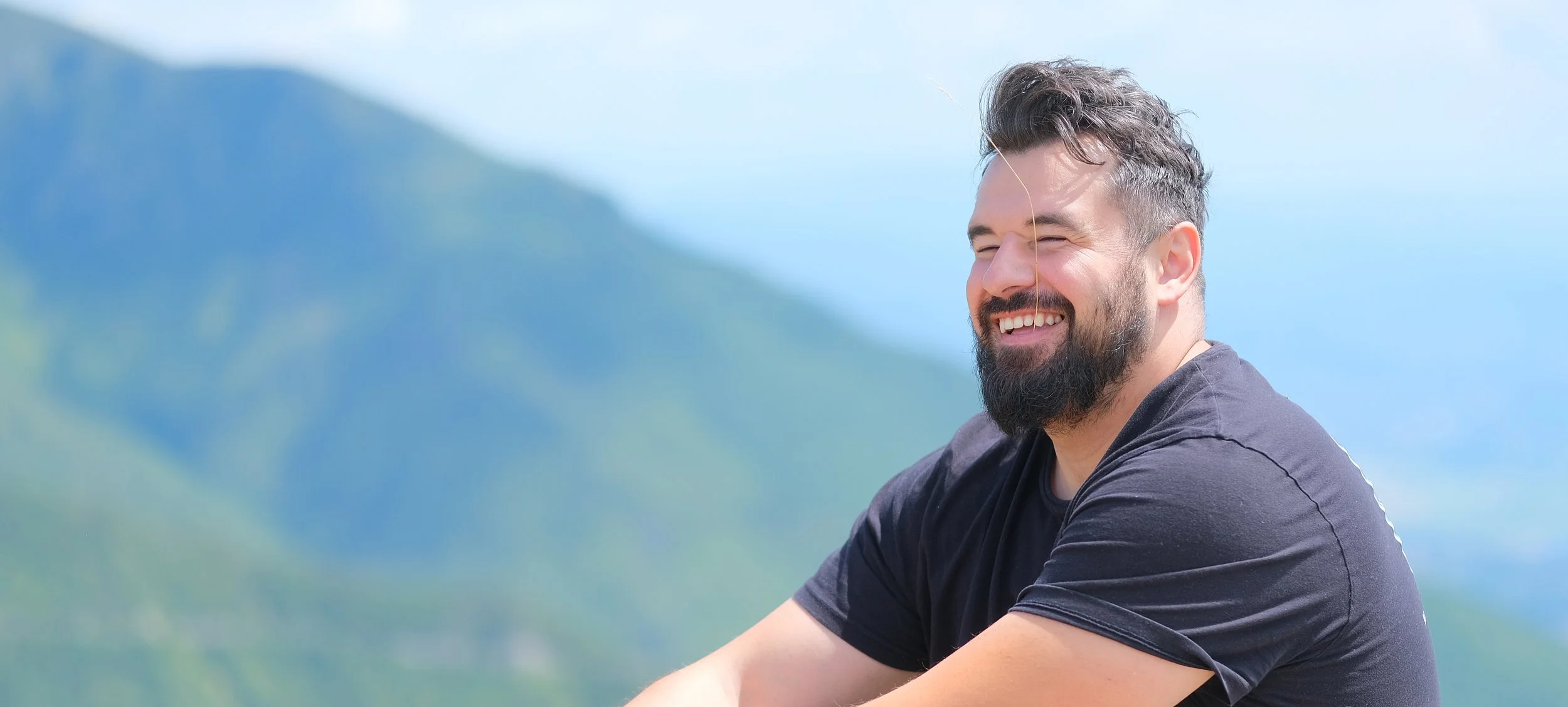 A smiling man with dark hair and a beard, wearing a black t-shirt, is outdoors with a blurred mountain and blue sky background.