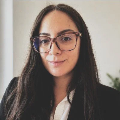 A woman with long dark hair, wearing glasses and a black blazer, smiling at the camera in a professional setting.