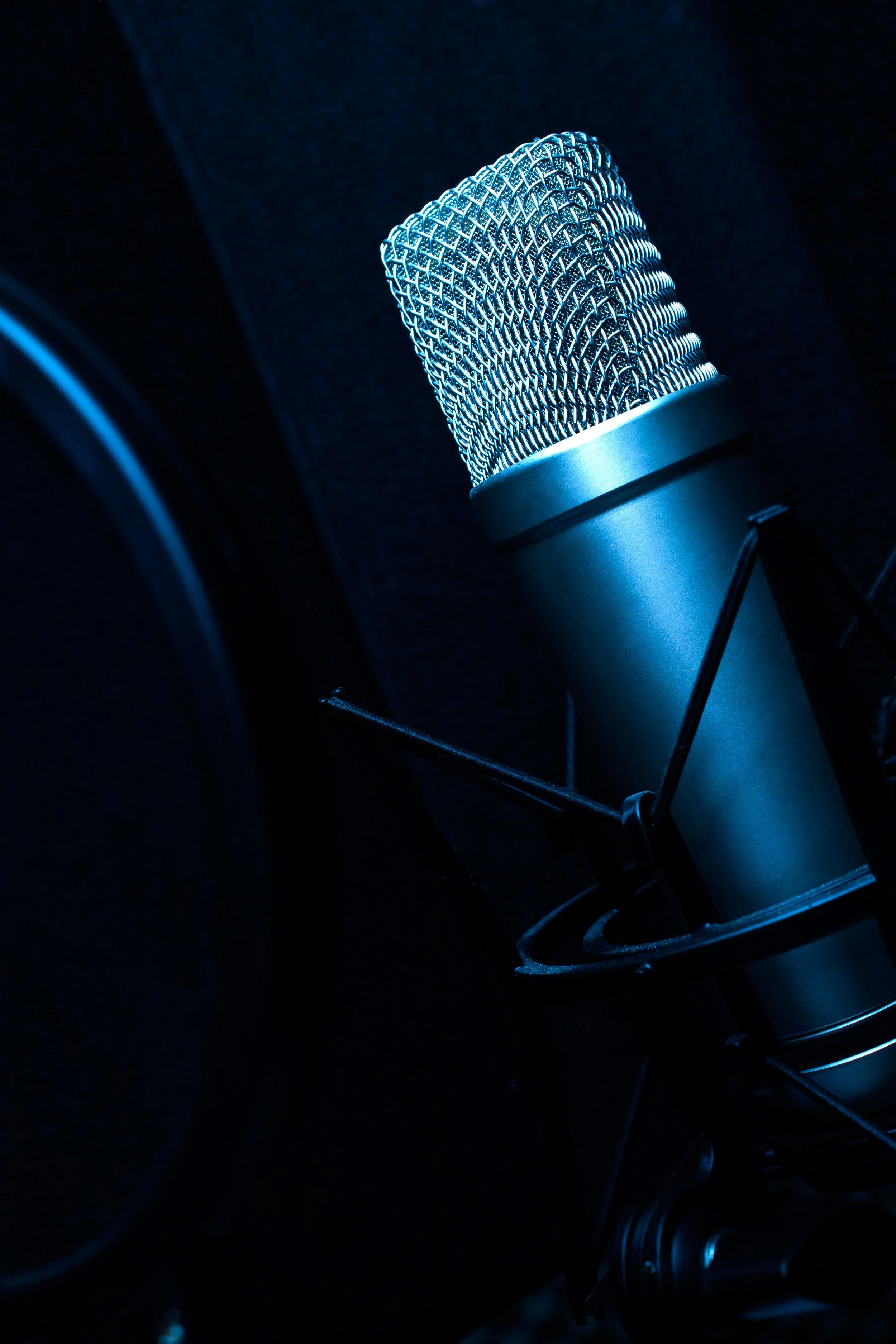 Close-up of a silver studio microphone with a mesh grille, illuminated by blue light, against a dark background.