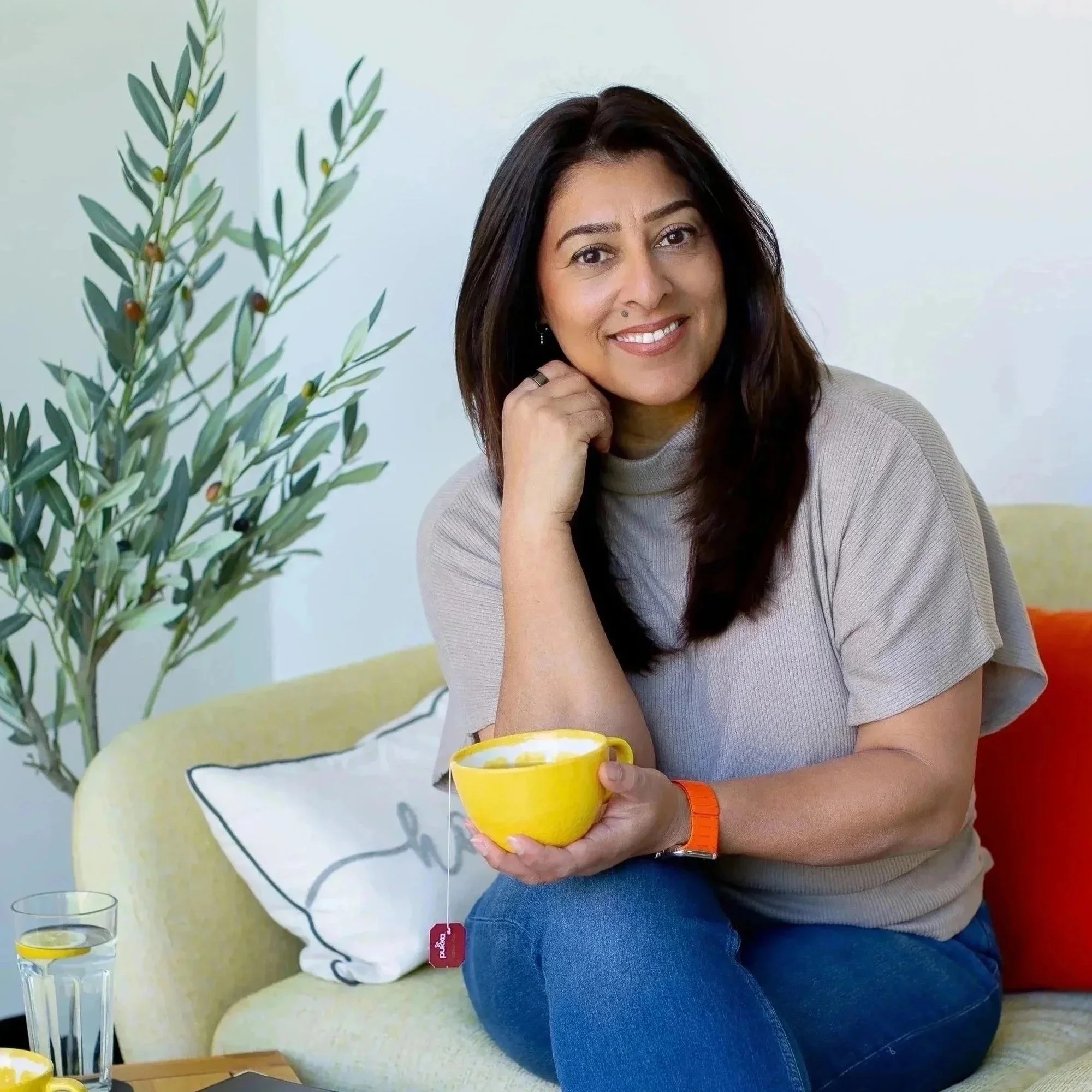 Picture of health coaching, smiling and looking at camera, sitting on sofa with cup of tea in one hand.
