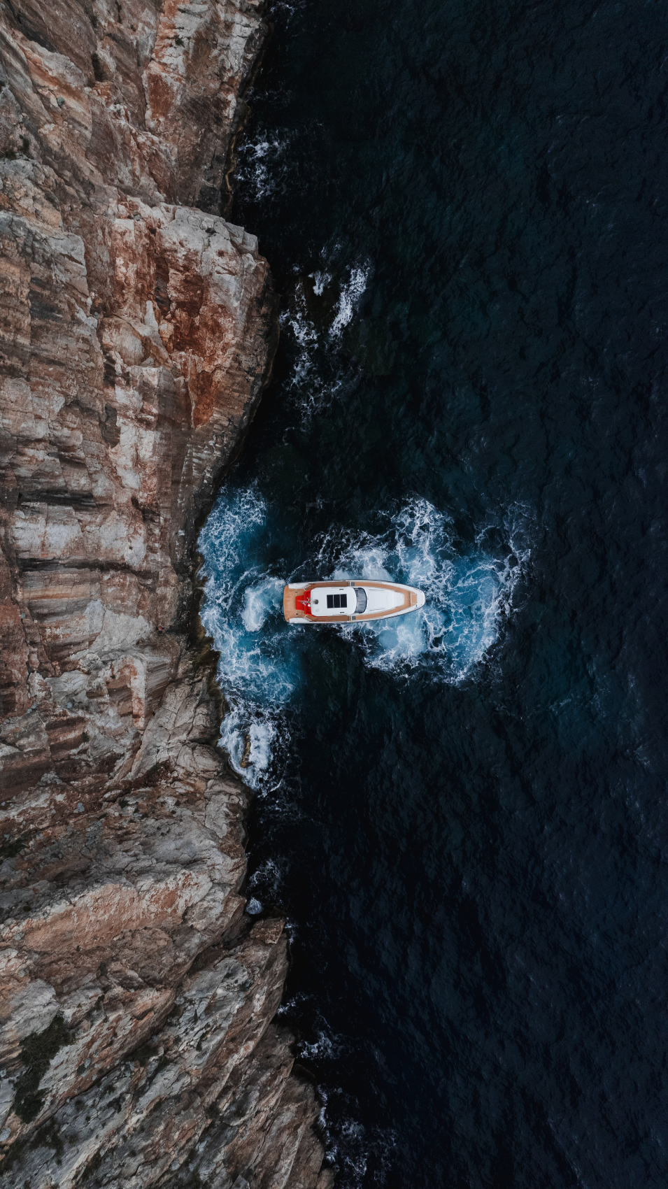 Overhead view of a boat traveling near a rocky cliff on the ocean, creating waves in the water.