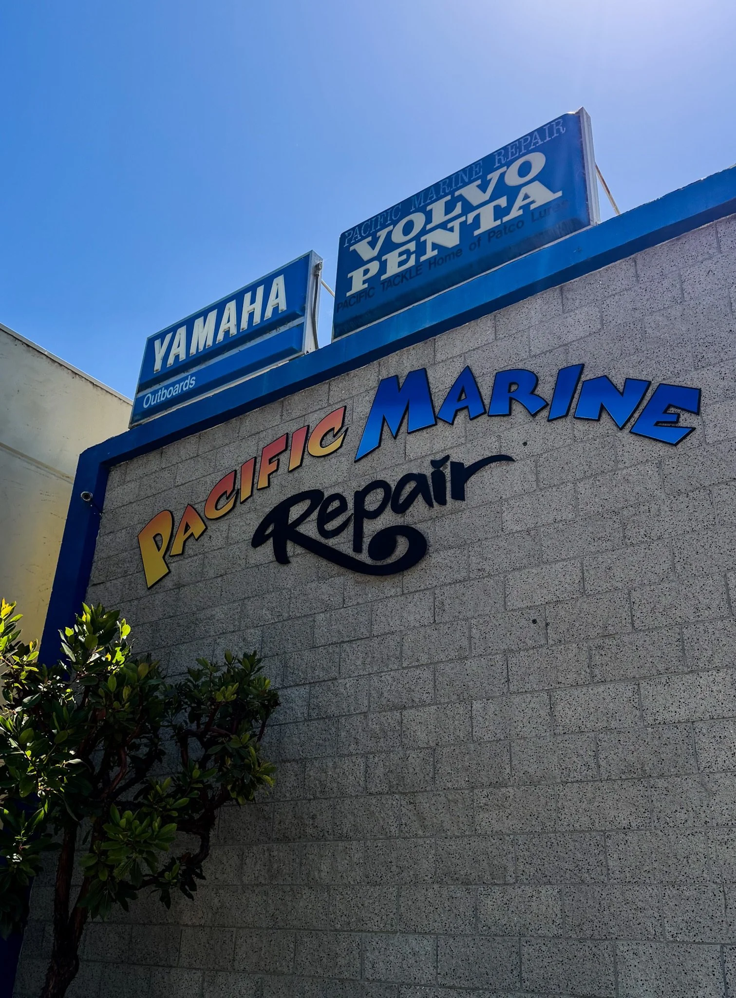 Sign on a building with blue background and white text for Yamaha Outboards, and another sign indicating Pacific Marine Repair with text in blue and yellow, along with a partial sign for Vovo Penta, also in blue and white.