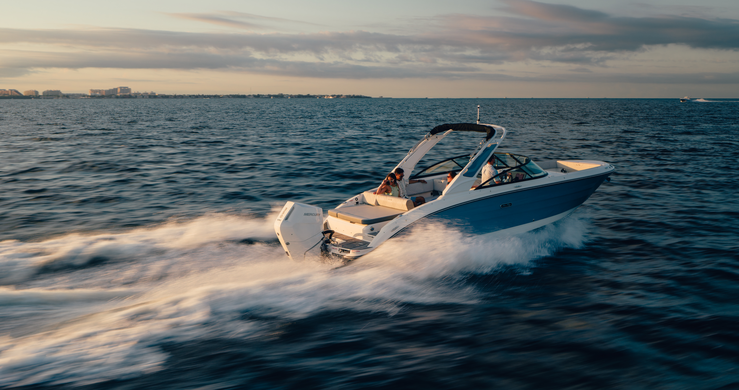 A white motorboat speeding across a body of water with four people onboard, surrounded by open water and a distant cityscape under a partly cloudy sky in the evening.