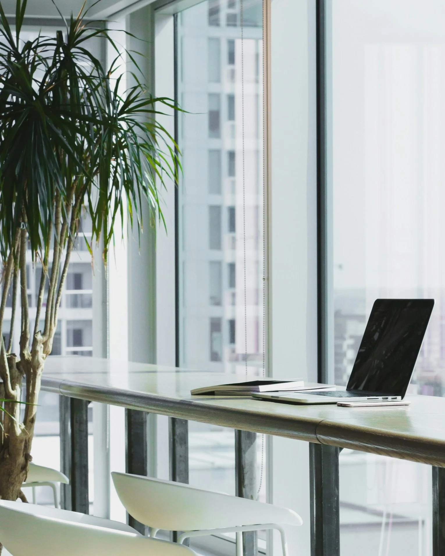 Modern office with a wooden desk, a laptop, an open notebook, a potted plant near large window with city view, and white chairs.