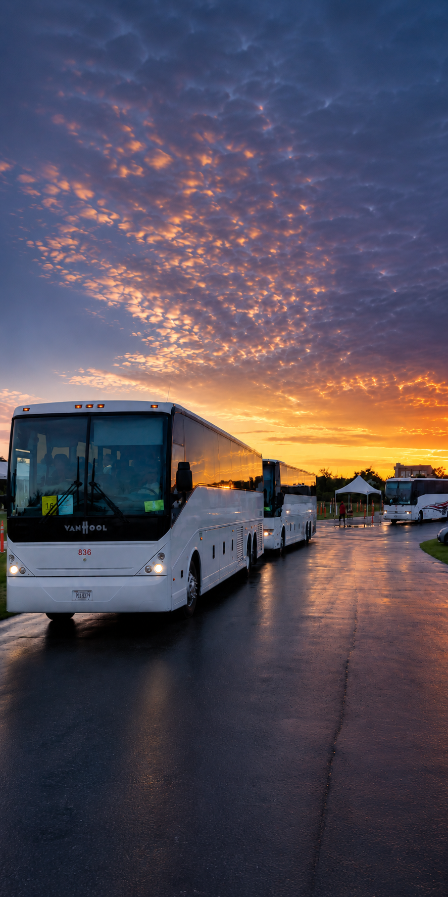 White tour buses parked on wet pavement during sunset with dramatic orange and blue sky and scattered clouds in the background.