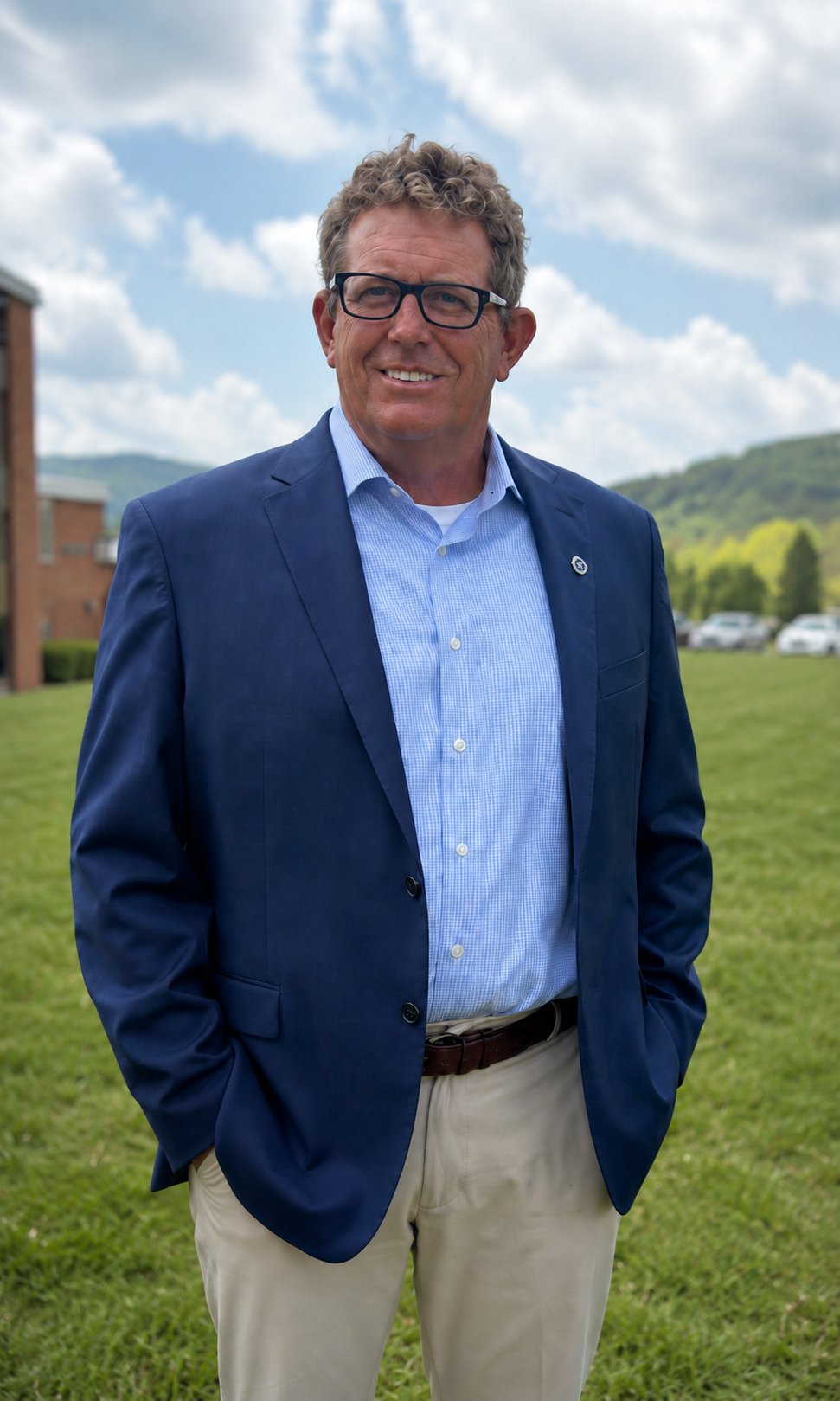 A middle-aged man with curly hair and glasses standing outdoors on a grassy area, smiling, wearing a blue suit jacket, light blue shirt, and khaki pants, with a background of trees, cars, and a partly cloudy sky.