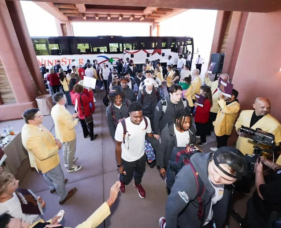 People arriving at a bus terminal, some wearing yellow jackets, others with backpacks, under a covered area with a bus in the background.