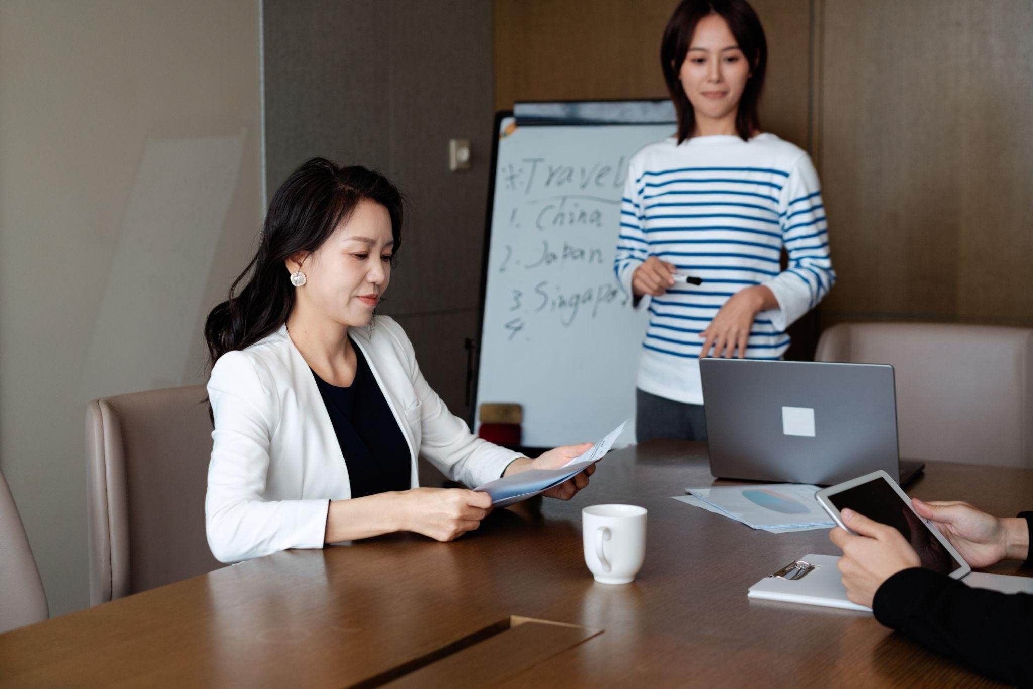Two women are in a meeting room with a laptop, papers, and a whiteboard. The woman in the foreground is looking at documents, wearing a white blazer, and sitting in a beige chair. The woman in the background, standing near the whiteboard with handwritten notes about travel destinations, wears a striped shirt and looks down at her hand.