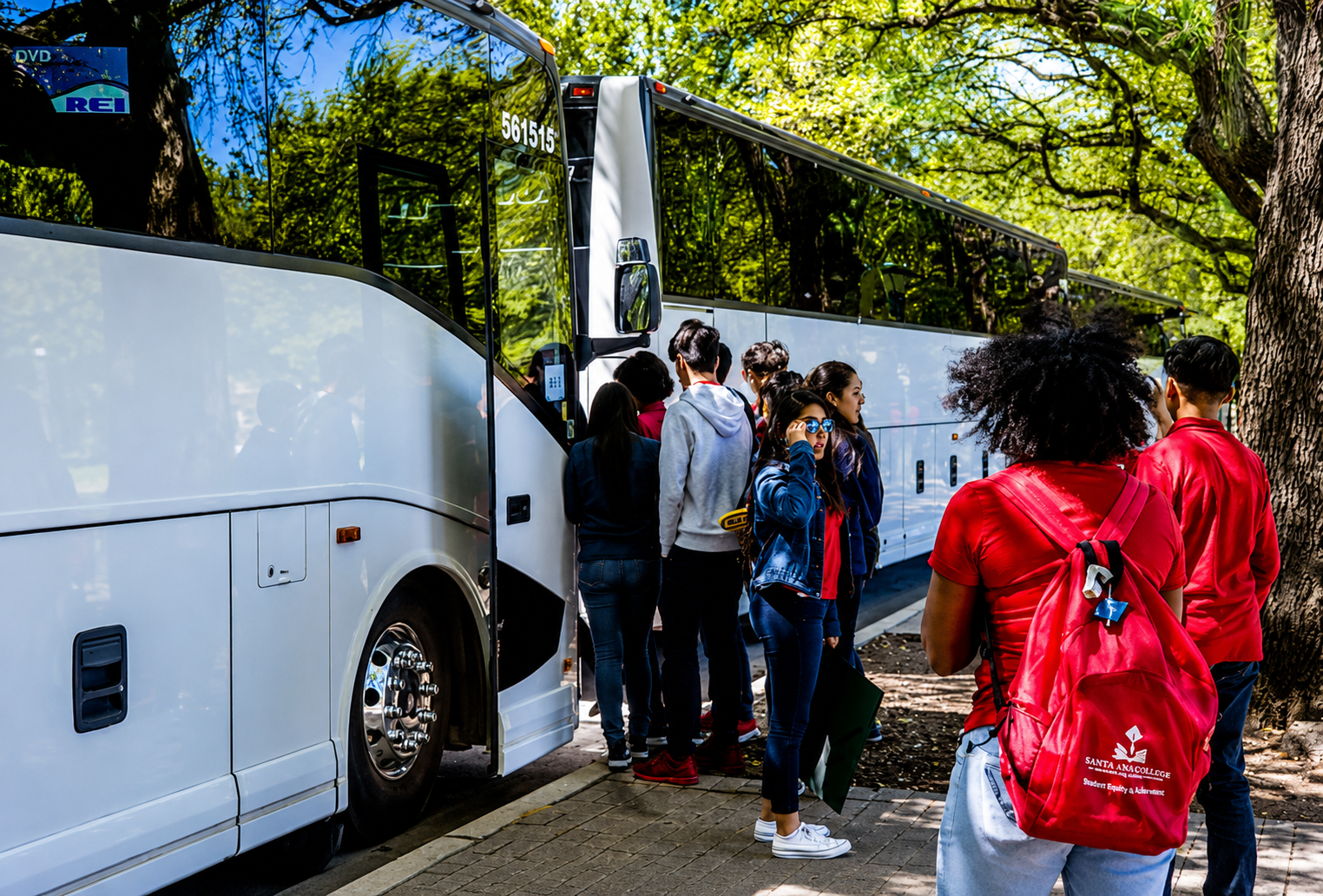 A group of people lined up to board a white tour bus parked beside a sidewalk under green trees on a sunny day.