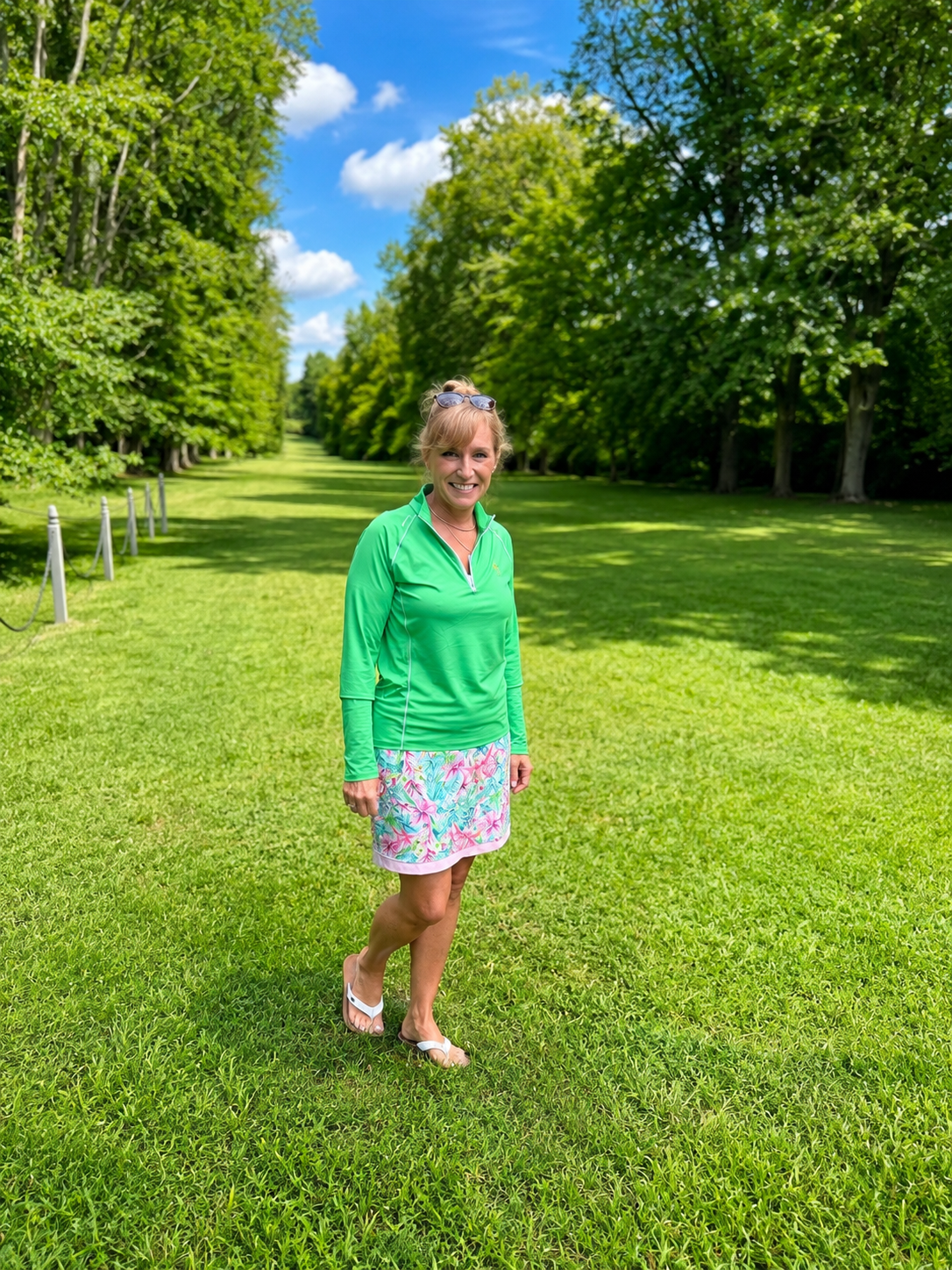 A woman in a green swim shirt and floral skirt stands on grass in a park with trees and blue sky in the background.