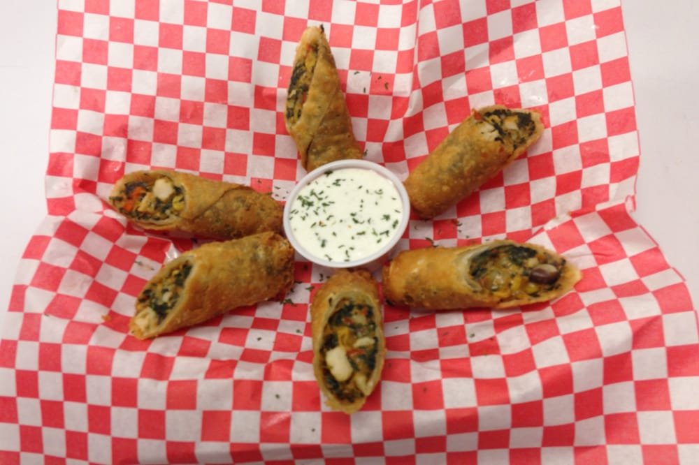 Six fried spring rolls arranged in a circle around a cup of white dipping sauce with herbs, on red and white checkered paper.