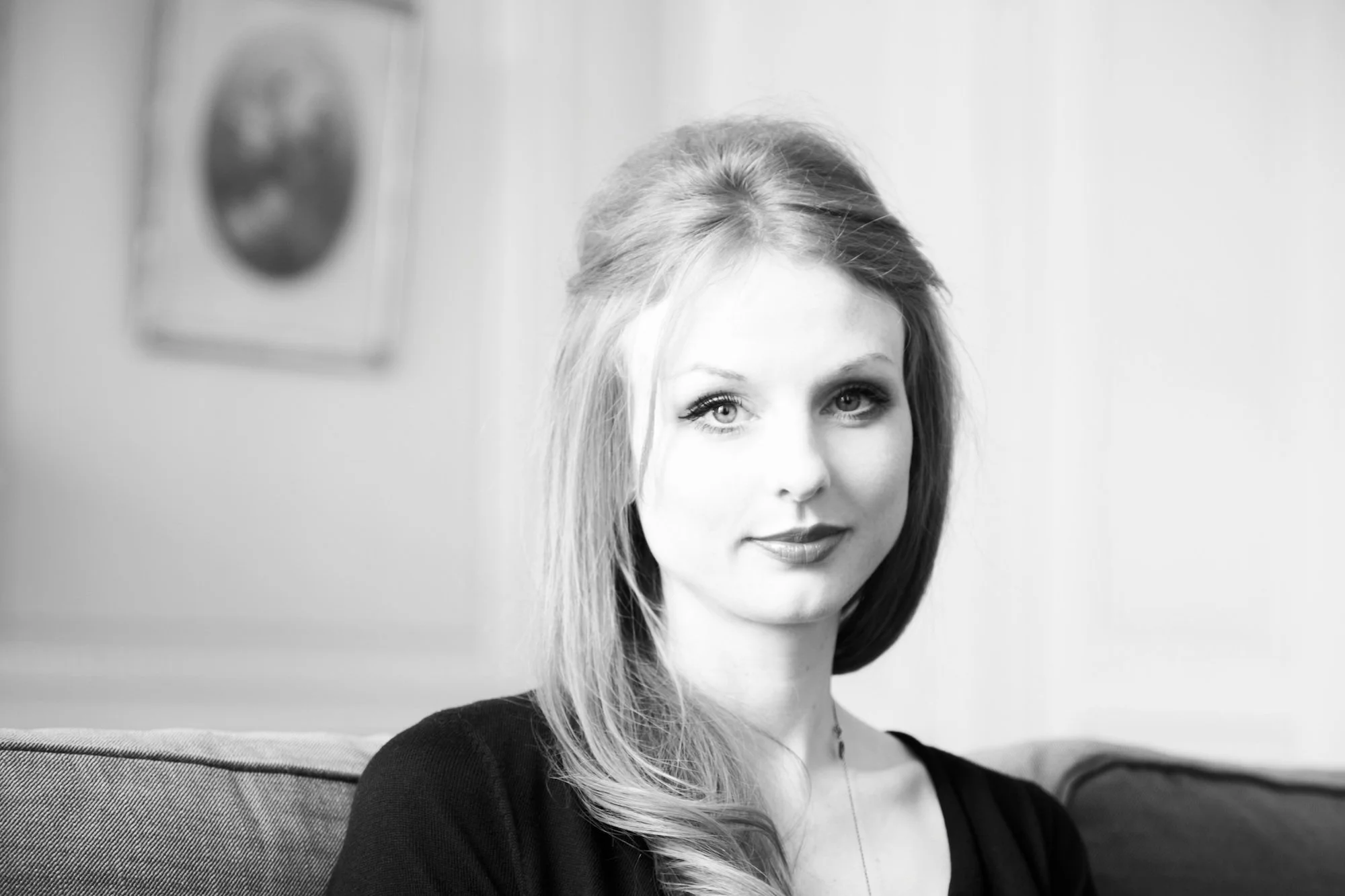 A black-and-white photograph of a young woman with long hair, sitting on a couch with a slight smile. She is wearing a dark top and is in a bright room with a blurred painting on the wall in the background.