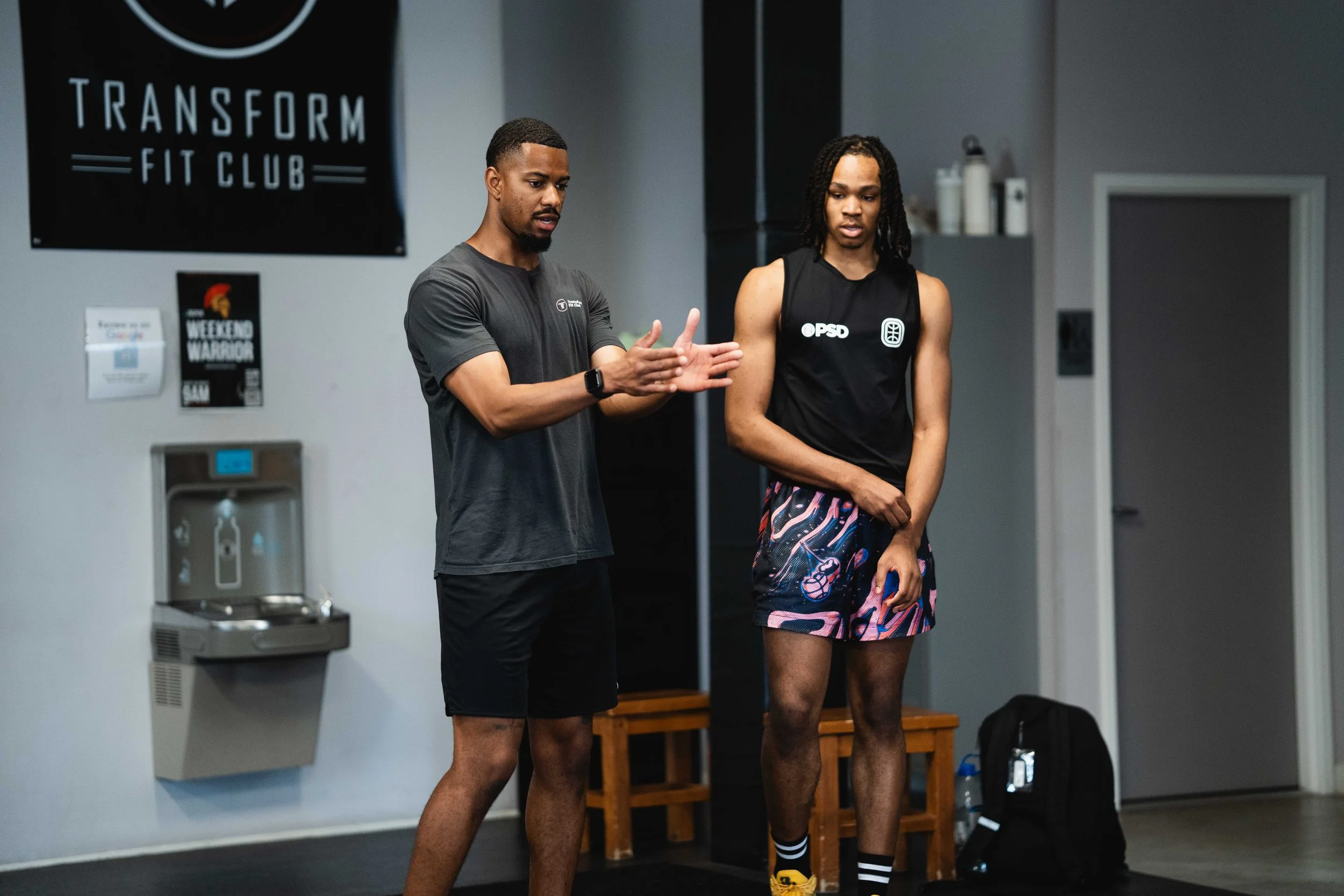 Two young men in athletic clothing are inside a gym. One is demonstrating an exercise while the other observes. The gym has a water dispenser, posters on the wall, and a wooden bench.