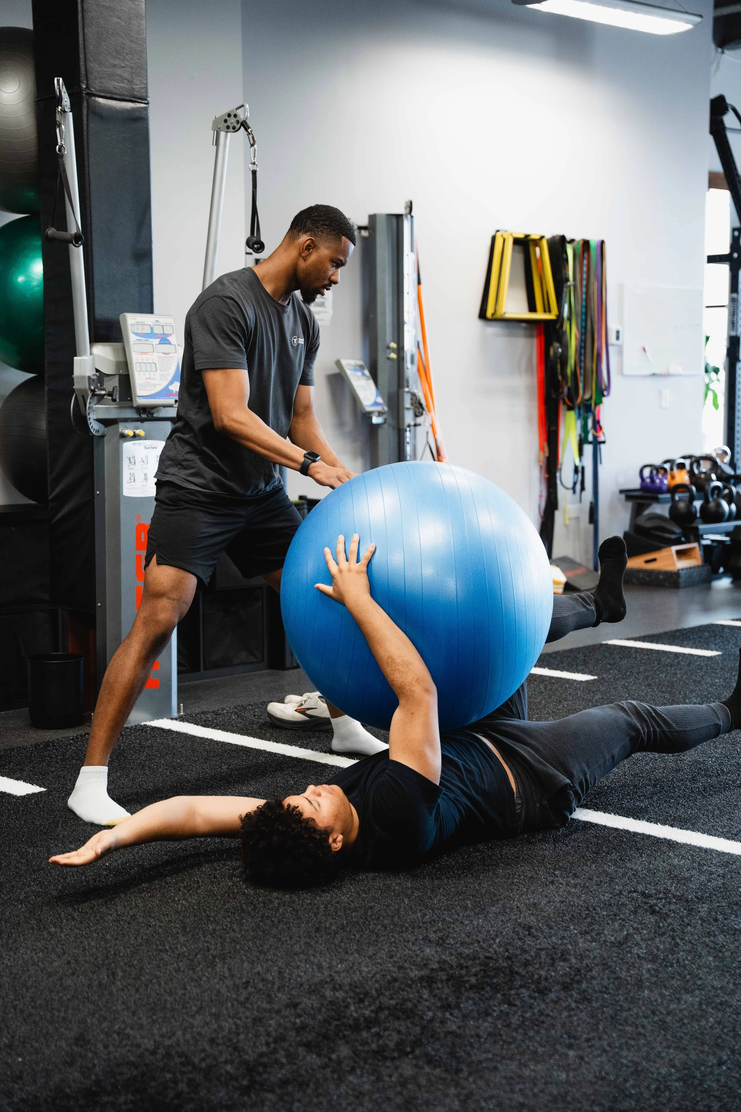A personal trainer is assisting a woman with physical therapy exercises using a large exercise ball. The woman is lying on her back on a black gym mat, with her arm raised and extended, while the trainer is standing beside her, holding the ball. The 