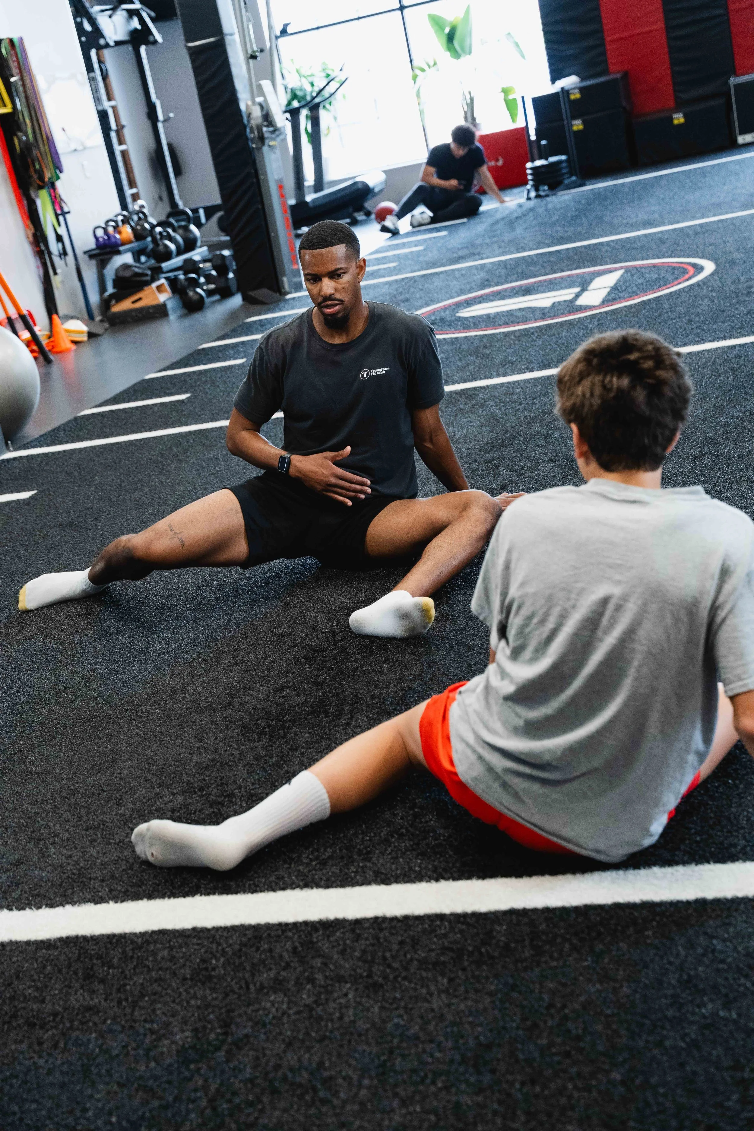 A trainer instructs a young boy during a stretching exercise at a gym with various fitness equipment in the background.