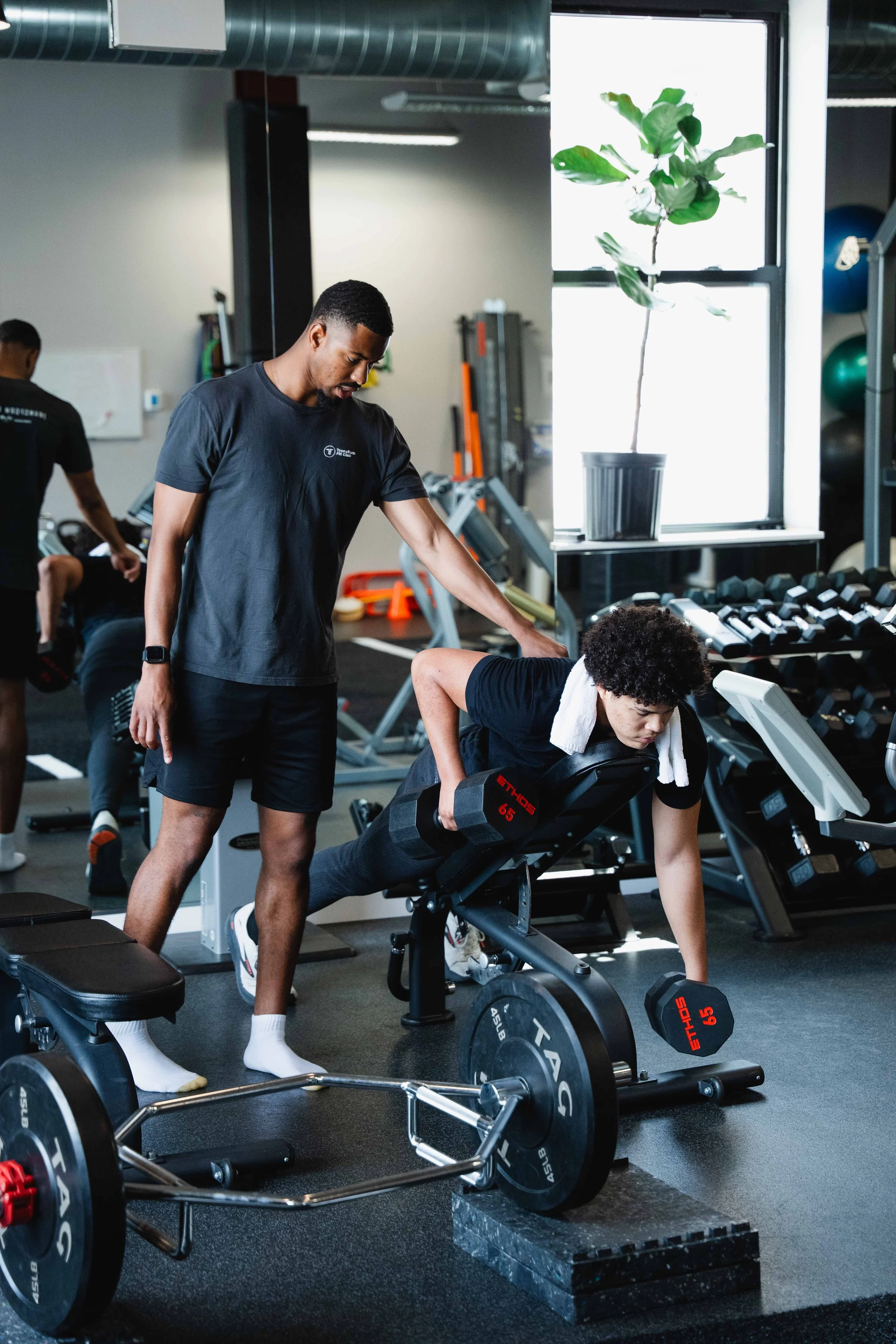 Personal trainer in black t-shirt assisting a woman with a dumbbell exercise in a gym, with fitness equipment and large windows in the background.